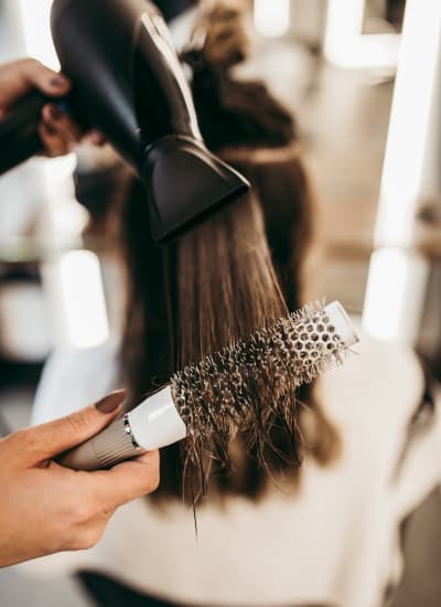 Hair stylist using a blow dryer and round brush to style weft hair extensions, highlighting salon service for maintaining polished extensions.
