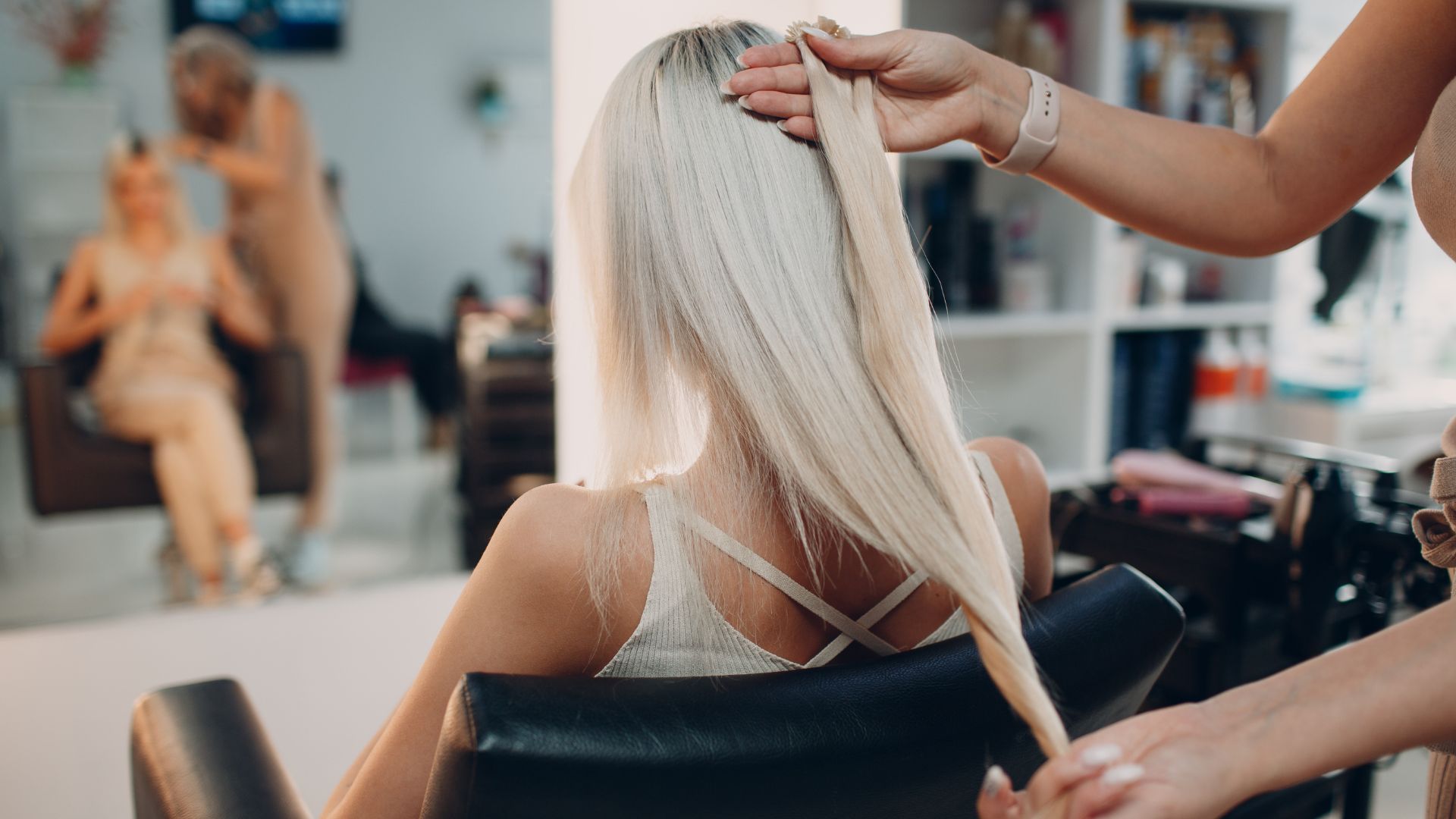 Woman in salon chair receiving K-tip hair extension application, stylist holding extension strand, showcasing hair enhancement process for volume and length.